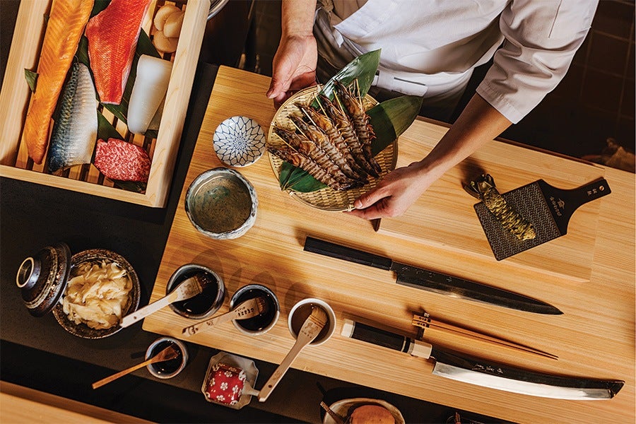 A person in a white chef's coat holds a woven basket with several large grilled shrimp on green leaves. The wooden table in front of them has various items including a long knife, a smaller knife with a sheath, chopsticks, a paddle with wasabi, small bowls with sauces, a container with pickled ginger, and a wooden box containing assorted raw fish and seafood.