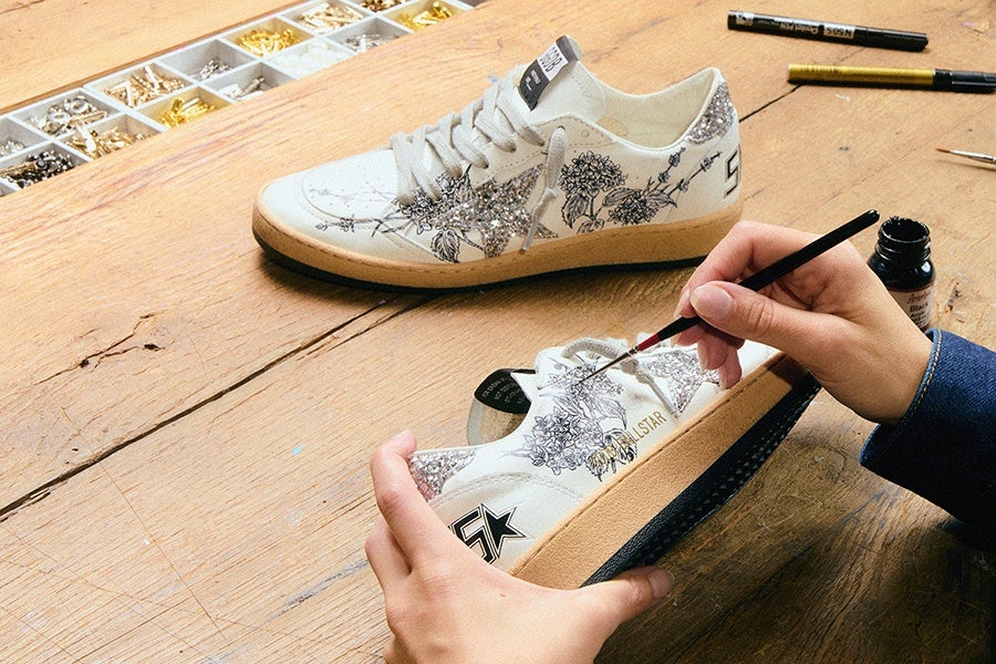 A pair of white sneakers with beige soles are being hand-painted with intricate black floral designs. One sneaker is placed on a wooden surface, while the other is held in hands with a fine paintbrush applying details. The sneakers feature glittery silver accents and a black star logo. Nearby, there are containers with small gold and silver decorative pieces and two paint markers on the table.