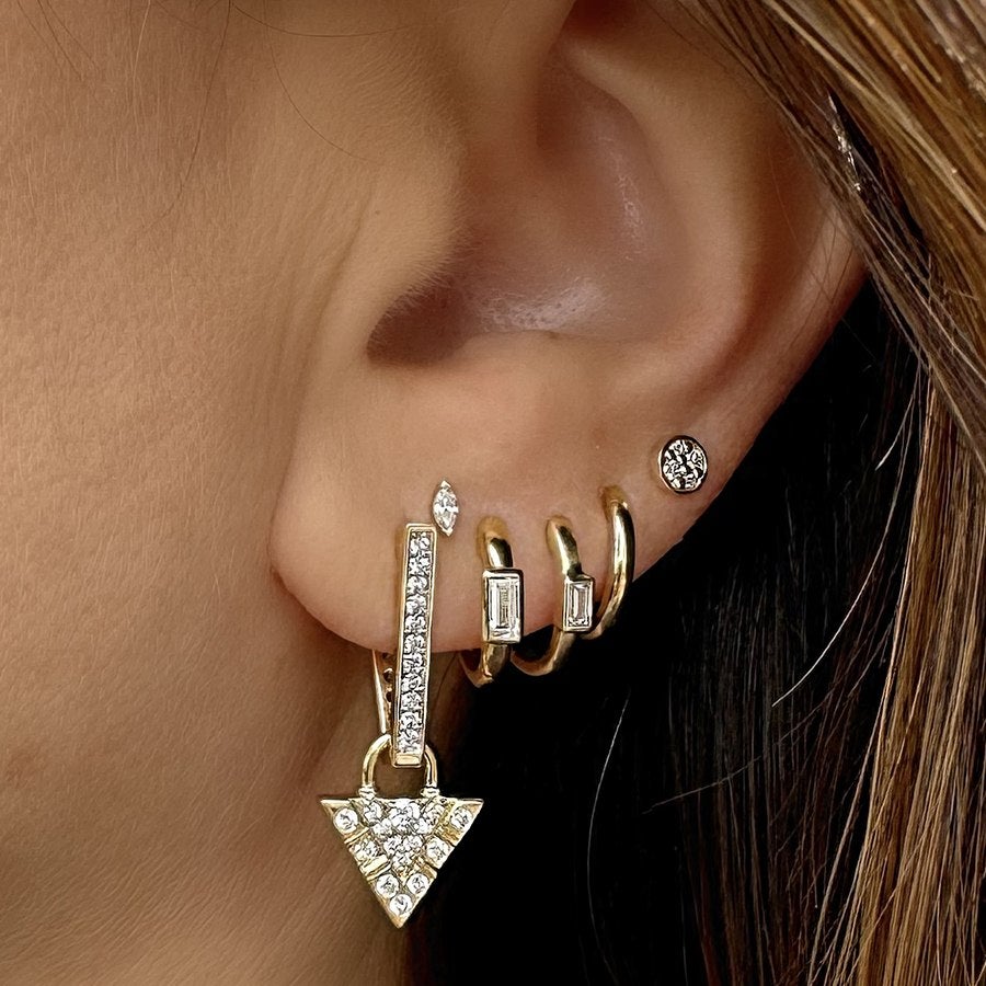 Close-up of a left ear adorned with five gold earrings: a long rectangular drop earring with embedded clear stones and a triangular pendant, two small gold hoops each with a rectangular clear stone, a plain gold hoop, and a small round stud with a floral pattern.