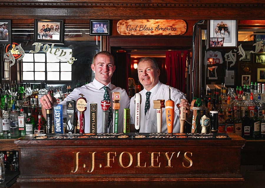 Two bartenders in white shirts and patterned ties stand behind a wooden bar with multiple beer taps. The bar front is engraved with "J.J. FOLEY'S." Above the bartenders, a wooden sign reads "God Bless America." The background shows shelves stocked with various bottles of liquor and framed photos on the walls.