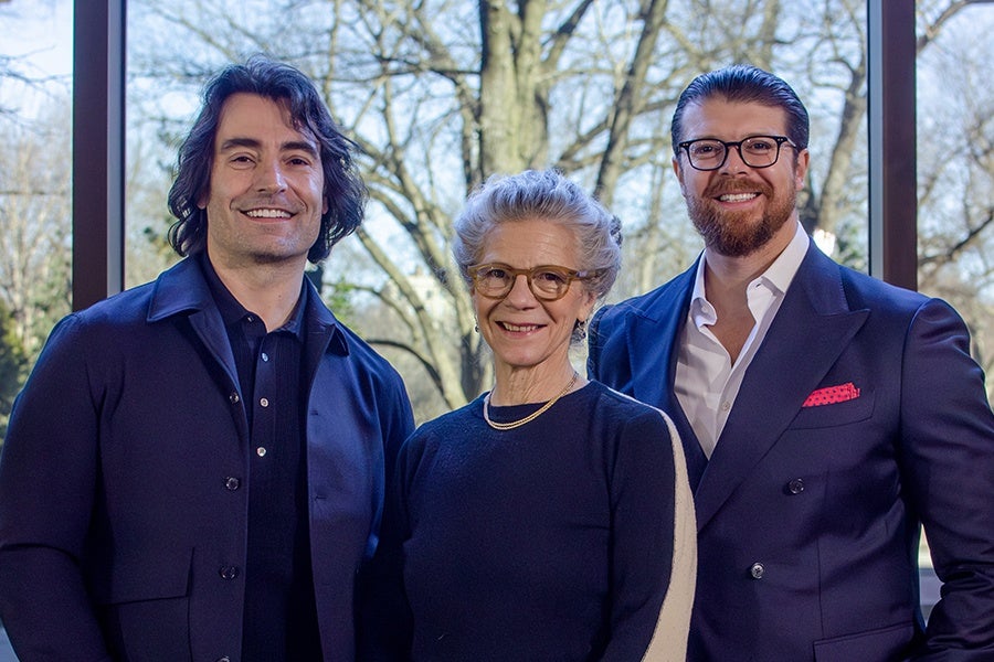 Three people standing indoors in front of large windows with a view of leafless trees outside. The person on the left has medium-length dark hair and is wearing a dark jacket over a dark shirt. The person in the middle has gray hair pulled back, glasses, and is wearing a dark sweater with a gold necklace. The person on the right has dark hair slicked back, a beard, glasses, and is wearing a dark suit with a white shirt and a red pocket square. All three are smiling.