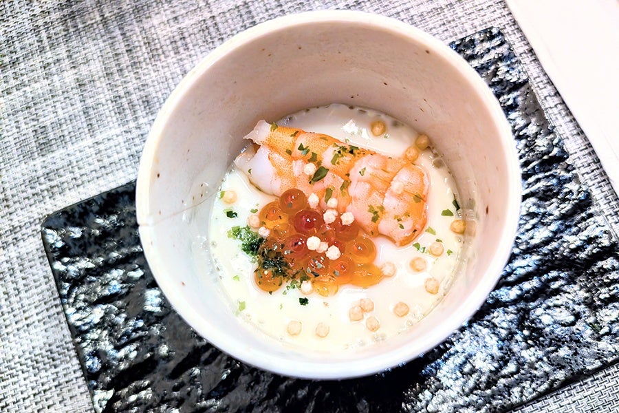 A white ceramic cup containing a creamy dish topped with a cooked shrimp, orange fish roe, small crunchy bits, and green herbs. The cup is placed on a textured black rectangular plate, which rests on a woven gray placemat.