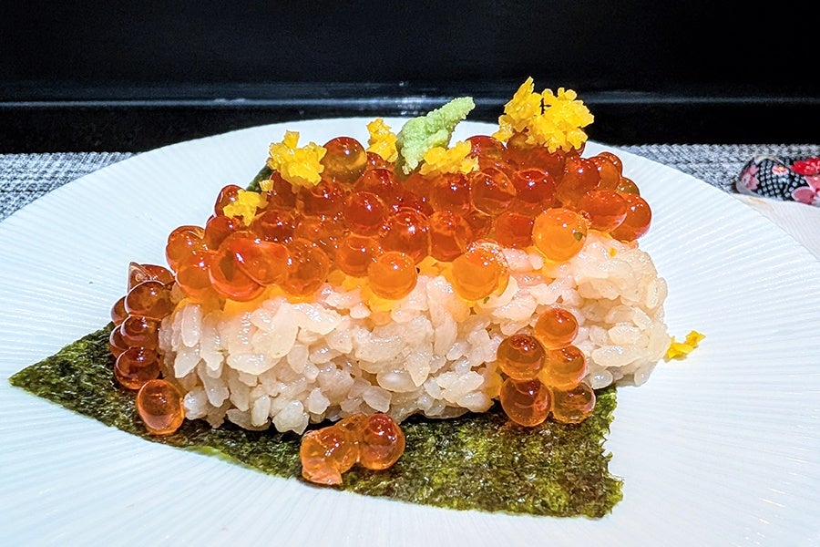 A piece of sushi featuring a rectangular block of rice topped with bright orange salmon roe, small yellow flower petals, and a small dollop of green wasabi, all resting on a square sheet of dark green seaweed on a white plate.