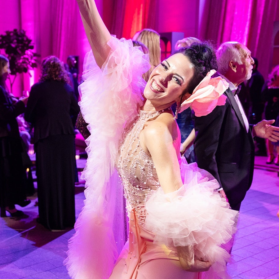 A joyful woman dressed in a sparkling, light pink gown with intricate beadwork and voluminous tulle sleeves is dancing at an event. She has a large pink flower accessory in her dark hair and is smiling brightly. The background shows other guests in formal attire under vibrant pink and purple lighting.