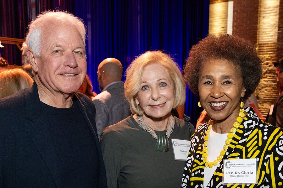 Three people smiling at an indoor event: an older man with white hair wearing a dark blazer and shirt, a woman with short blonde hair wearing a dark top and a necklace, and a woman with short curly hair wearing a patterned yellow, black, and white jacket with a yellow beaded necklace. The two women have name tags, one reading "Susan" and the other "Rev. Dr. Gloria White-Hammond." The background features blue curtains and warm lighting.