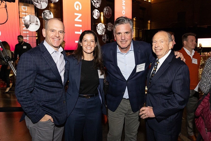 Four people dressed in business attire stand closely together, smiling at the camera. The background features large red banners with white text and circular black-and-white photos, suggesting a formal event or conference setting. The lighting is warm and the atmosphere appears lively.