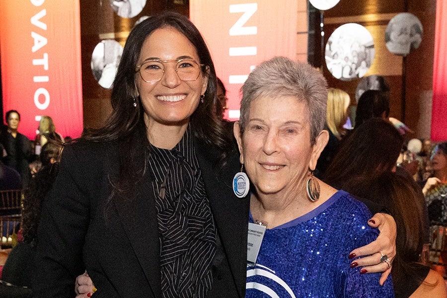 Two women smiling and posing together at an indoor event. The woman on the left has long dark hair, wears glasses, a black blazer, and a patterned black blouse. The woman on the right has short gray hair, wears large earrings, and a sparkly blue top with a circular design. The background shows other people seated and red banners with white text.