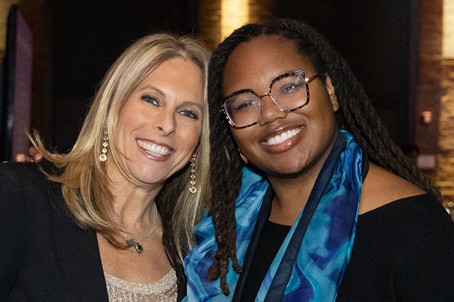 Two women smiling closely together; the woman on the left has blonde hair, wears a black blazer, gold earrings, and a sparkly top, while the woman on the right has long dark dreadlocks, glasses, a blue patterned scarf, and a black top.