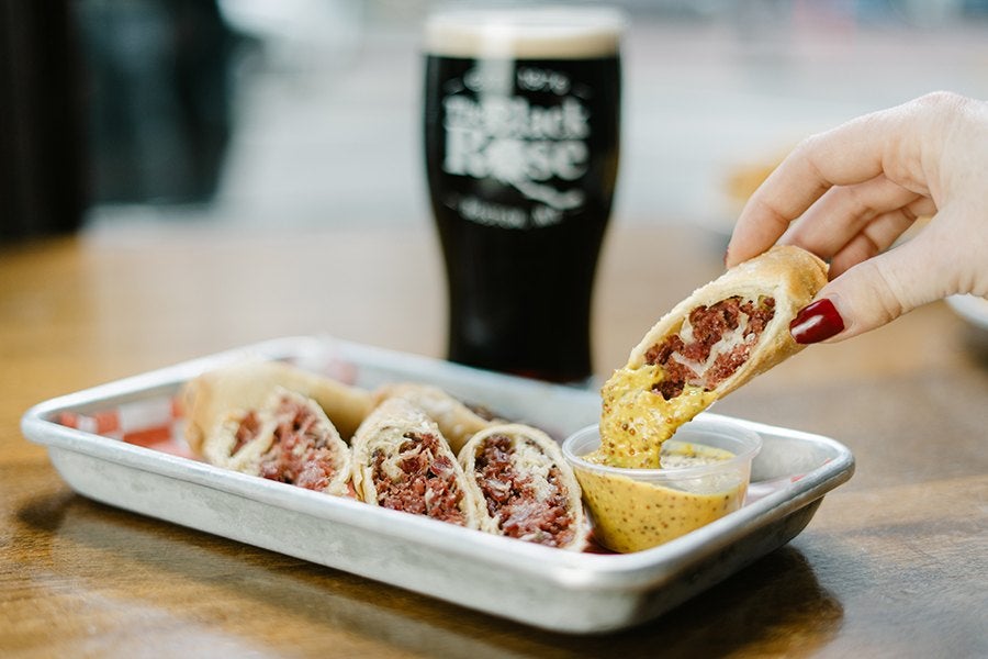 A metal tray holds several pieces of a rolled sandwich filled with corned beef or pastrami. One piece is being dipped into a small plastic cup of grainy mustard by a hand with red-painted nails. In the background, there is a dark glass of beer with a creamy head, slightly out of focus.