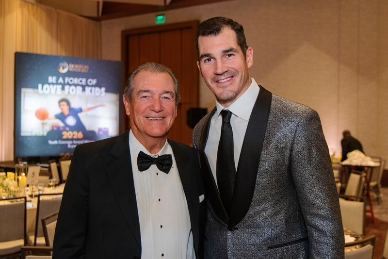 Two men dressed in formal attire stand side by side in a banquet hall. The man on the left wears a black tuxedo with a white shirt and black bow tie, while the man on the right wears a silver patterned tuxedo jacket with black lapels, a white shirt, and a black tie. In the background, there is a large screen displaying the text "BE A FORCE OF LOVE FOR KIDS" and "2026 Youth Orange Awards." The setting includes tables and chairs arranged for a formal event.