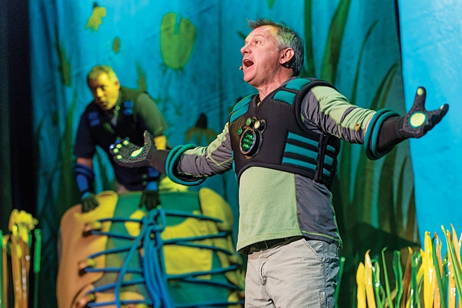 A man in a green and gray long-sleeve shirt and beige pants is performing on stage, wearing a black and teal vest with circular green lights and matching gloves. Another person in a dark outfit is in the background, partially obscured, near a large prop resembling a colorful insect or creature. The backdrop features blue and green tones with abstract plant-like shapes.