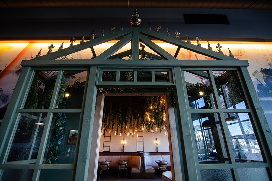 A decorative wooden greenhouse-style frame with glass panels and a peaked roof, adorned with small ornamental finials along the top edge. Inside, a cozy dining area is visible with hanging greenery and warm Edison-style light bulbs, wooden chairs, and cushioned bench seating against a white wall with ladder-like wall decorations. The background includes a mural and large windows reflecting outdoor scenery.