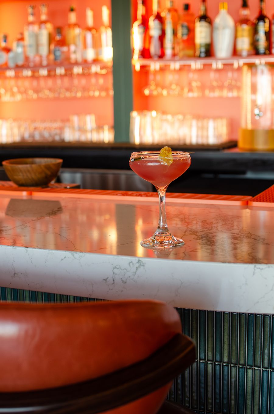 A pink cocktail in a coupe glass garnished with a small yellow bit of honeycomb, placed on a white marble bar counter. The background shows a blurred bar with bottles and glasses on orange-lit shelves. A brown leather bar stool is partially visible in the foreground.