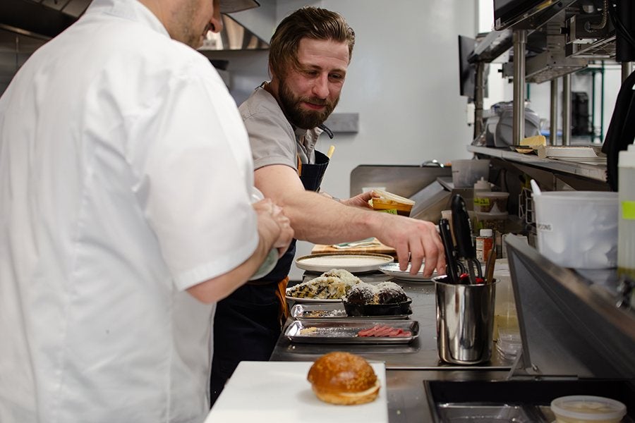 Two chefs are working in a professional kitchen, with one chef reaching for a container while the other watches. Various plated dishes and ingredients are arranged on the counter, including a sandwich bun on a cutting board in the foreground. Kitchen utensils are stored in a metal container nearby.