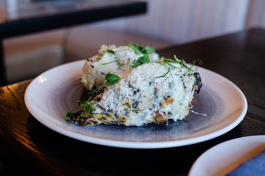 Two pieces of grilled cabbage topped with a creamy white sauce, grated cheese, and garnished with fresh green herbs, served on a white plate with a blue-gray center.