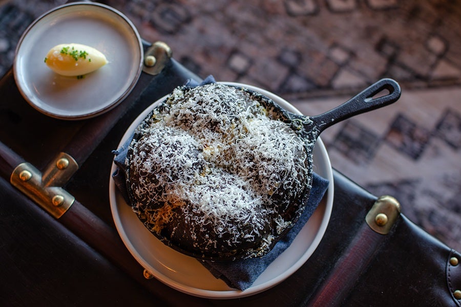 A cast iron skillet filled with dark bread rolls topped with grated cheese, placed on a white plate with a dark napkin underneath. Next to it, a small metal dish holds a dollop of butter garnished with chopped herbs. The setting is on a dark wooden surface with a patterned fabric in the background.