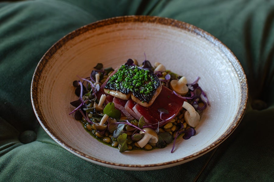 Seared fish fillet topped with chopped chives, served on a bed of thinly sliced red vegetables, mushrooms, microgreens, and a light sauce in a rustic, shallow beige bowl.
