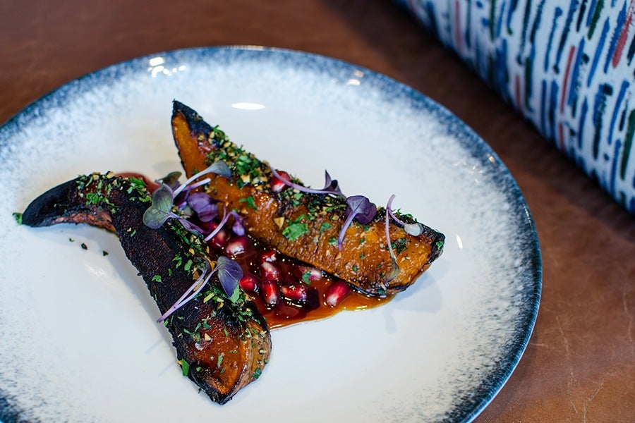 Two pieces of roasted squash garnished with chopped herbs and microgreens, served on a white plate with a blue-gray rim, accompanied by pomegranate seeds and a drizzle of sauce.