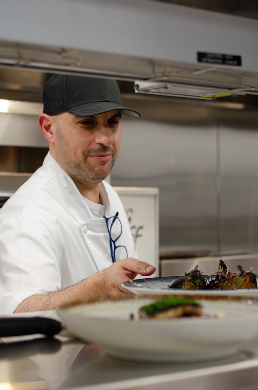 A chef wearing a white coat and black cap is holding a plate of food in a professional kitchen. The chef has blue glasses hanging from the coat and is focused on presenting the dish. The kitchen background is stainless steel, and there is another plate of food blurred in the foreground.