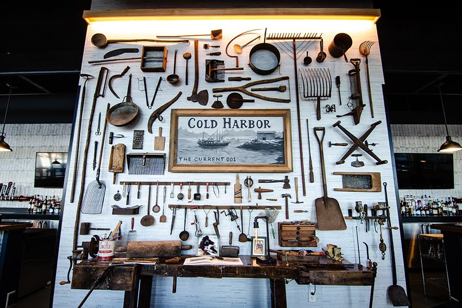 A wall display featuring a variety of vintage tools and utensils arranged around a framed sign that reads "Cold Harbor The Current 001." Below the display is a rustic wooden workbench with additional tools, a can labeled "Swift's," and an open book. The setting appears to be indoors with modern lighting and bar areas visible on either side.
