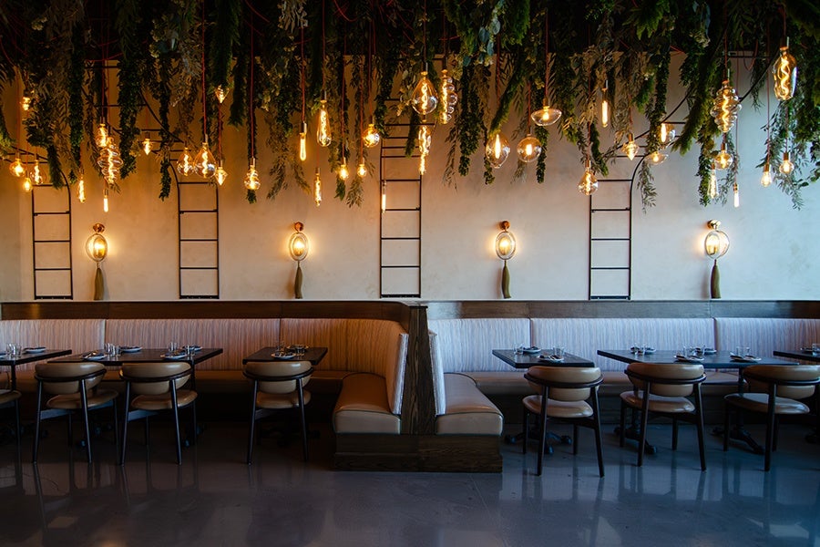 A modern restaurant interior featuring two long banquette seating areas separated by a wooden divider. The seating has light-colored upholstery with dark wooden frames. Above the seating, numerous hanging light bulbs with warm yellow light are suspended from the ceiling, interspersed with hanging green foliage. The walls are light-colored with decorative wall sconces and black ladder-like frames. Tables are set with plates, glasses, and cutlery, and the floor is a polished dark surface.