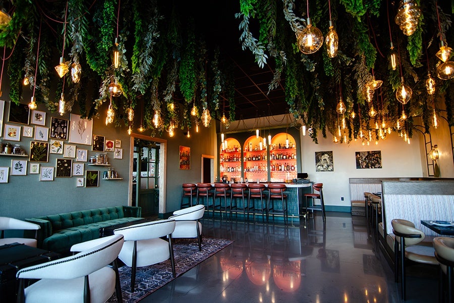 A stylish bar interior featuring a green tufted sofa, white cushioned chairs, and a dark wood bar with red bar stools. The ceiling is adorned with hanging greenery and various warm, decorative pendant lights. The walls display framed botanical artwork, and the floor has a polished, reflective surface with a patterned rug near the seating area.