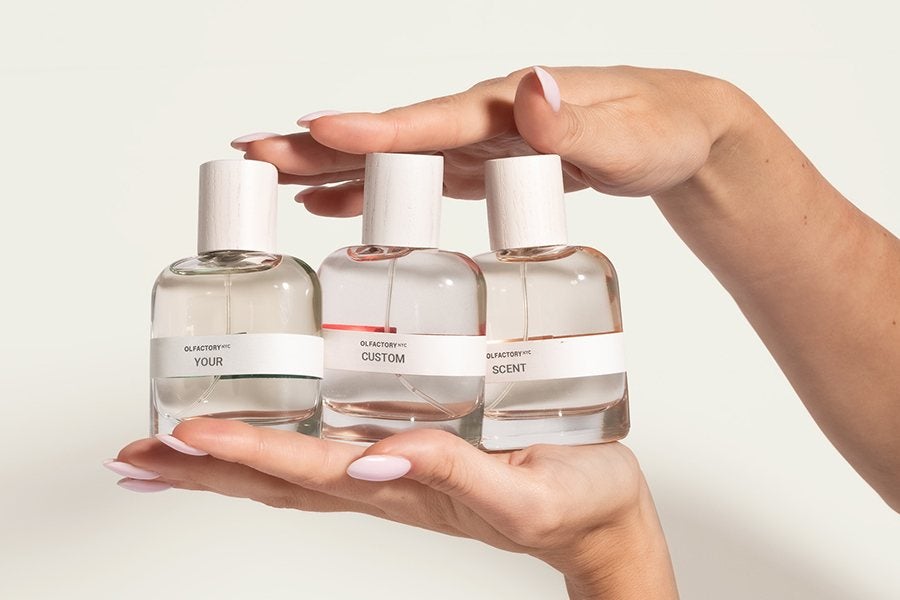 Three clear glass perfume bottles with white wooden caps are held between two hands with light pink manicured nails. Each bottle has a white label with black text reading "YOUR," "CUSTOM," and "SCENT" respectively, indicating a customizable fragrance set. The background is plain and light-colored.
