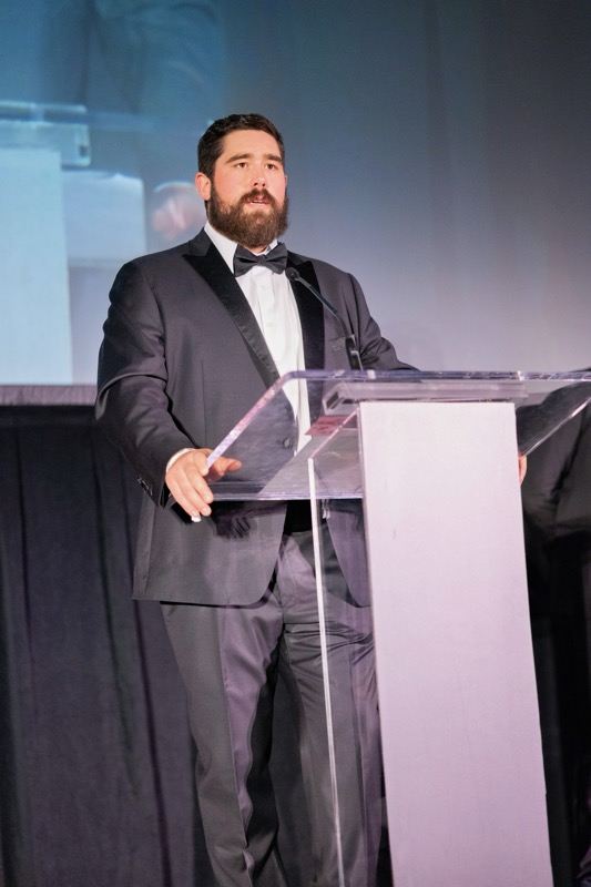 A man with a beard wearing a black tuxedo with a white shirt and black bow tie stands behind a clear podium, speaking. The background is dark with a large screen partially visible behind him.