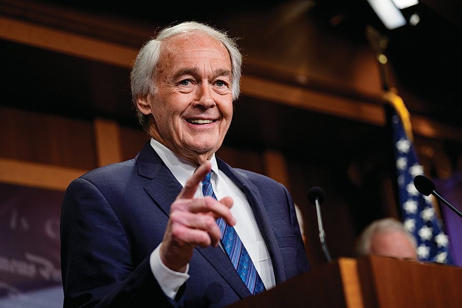An older man with white hair wearing a dark blue suit, white shirt, and blue patterned tie is standing behind a wooden podium, smiling and pointing forward. The background includes an American flag and blurred elements suggesting a formal setting.