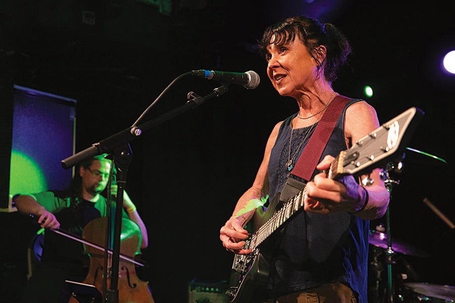 A woman with dark hair tied back is playing an electric guitar and singing into a microphone. She is wearing a sleeveless dark top and a necklace. In the background, a man is playing a cello, illuminated by green and purple stage lights. The setting appears to be a live music performance on a dimly lit stage.