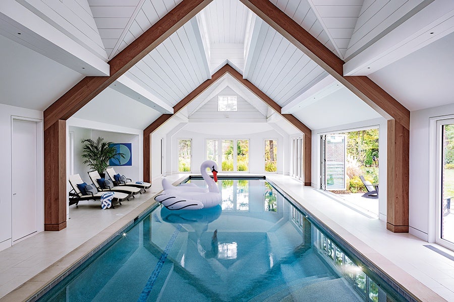 Indoor swimming pool with clear blue water, featuring a large inflatable swan float. The pool area has a high white ceiling with wooden beams and large windows letting in natural light. On one side, there are several lounge chairs with cushions and a potted plant. The space opens to an outdoor garden area visible through glass doors.