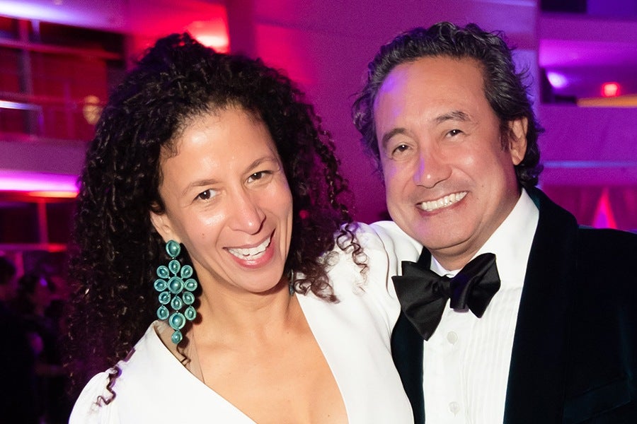 A smiling woman with curly dark hair wearing large turquoise earrings and a white outfit stands next to a smiling man in a black tuxedo with a white shirt and black bow tie. The background features purple and pink lighting.