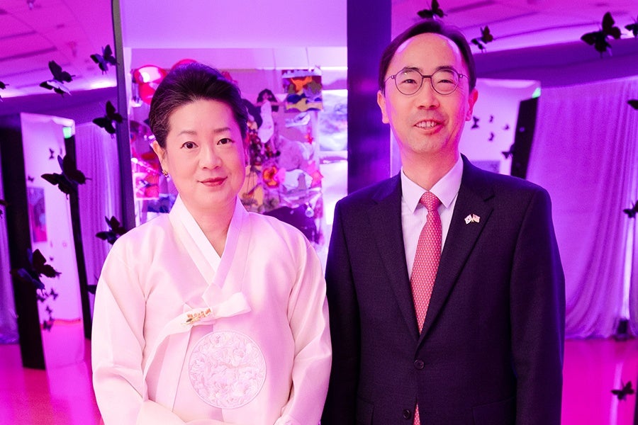 A woman wearing a traditional light pink hanbok with floral embroidery stands next to a man in a dark suit, white shirt, and red patterned tie. Both are smiling, and the background is illuminated with purple lighting and decorated with black butterfly silhouettes.