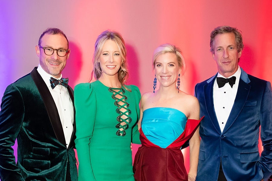 Two men and two women dressed in formal evening attire stand together against a gradient background of purple and red. The man on the left wears a dark green velvet tuxedo jacket with a white shirt and a patterned bow tie. The woman next to him wears a long-sleeved green dress with a lace-up front detail. The woman next to her wears a strapless dress with a blue bodice and a red skirt, accessorized with long blue earrings. The man on the right wears a dark blue velvet tuxedo jacket with a white shirt and a black bow tie. All four are smiling.