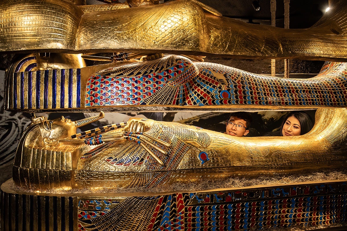 Two people are peeking out from inside an ornate, gold and colorful ancient Egyptian sarcophagus, which is decorated with intricate patterns and hieroglyphics. The sarcophagus is open, revealing the upper bodies of the individuals inside.