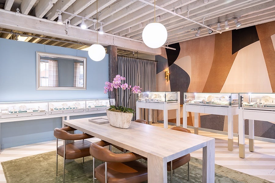 Modern jewelry store interior with a light wood table surrounded by brown leather chairs. A white planter with pink orchids is centered on the table. Display cases with jewelry line the walls, which feature a blue section with a large mirror and a geometric patterned wall in shades of brown, beige, and black. The ceiling has exposed beams and hanging round white pendant lights.