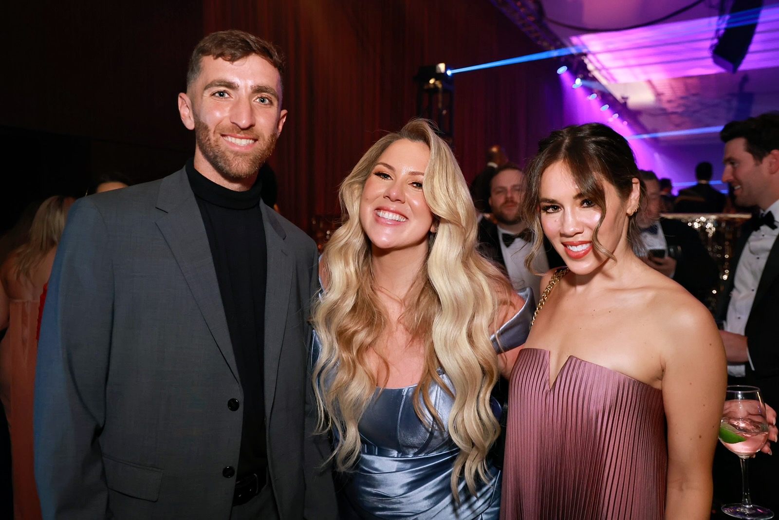 A man and two women smiling at a formal event. The man on the left is wearing a gray suit jacket over a black turtleneck. The woman in the center has long, wavy blonde hair and is wearing a shiny blue off-shoulder dress. The woman on the right has dark hair styled in loose curls and is wearing a strapless, pleated mauve dress, holding a glass with a lime wedge. The background features other guests in formal attire and purple lighting.