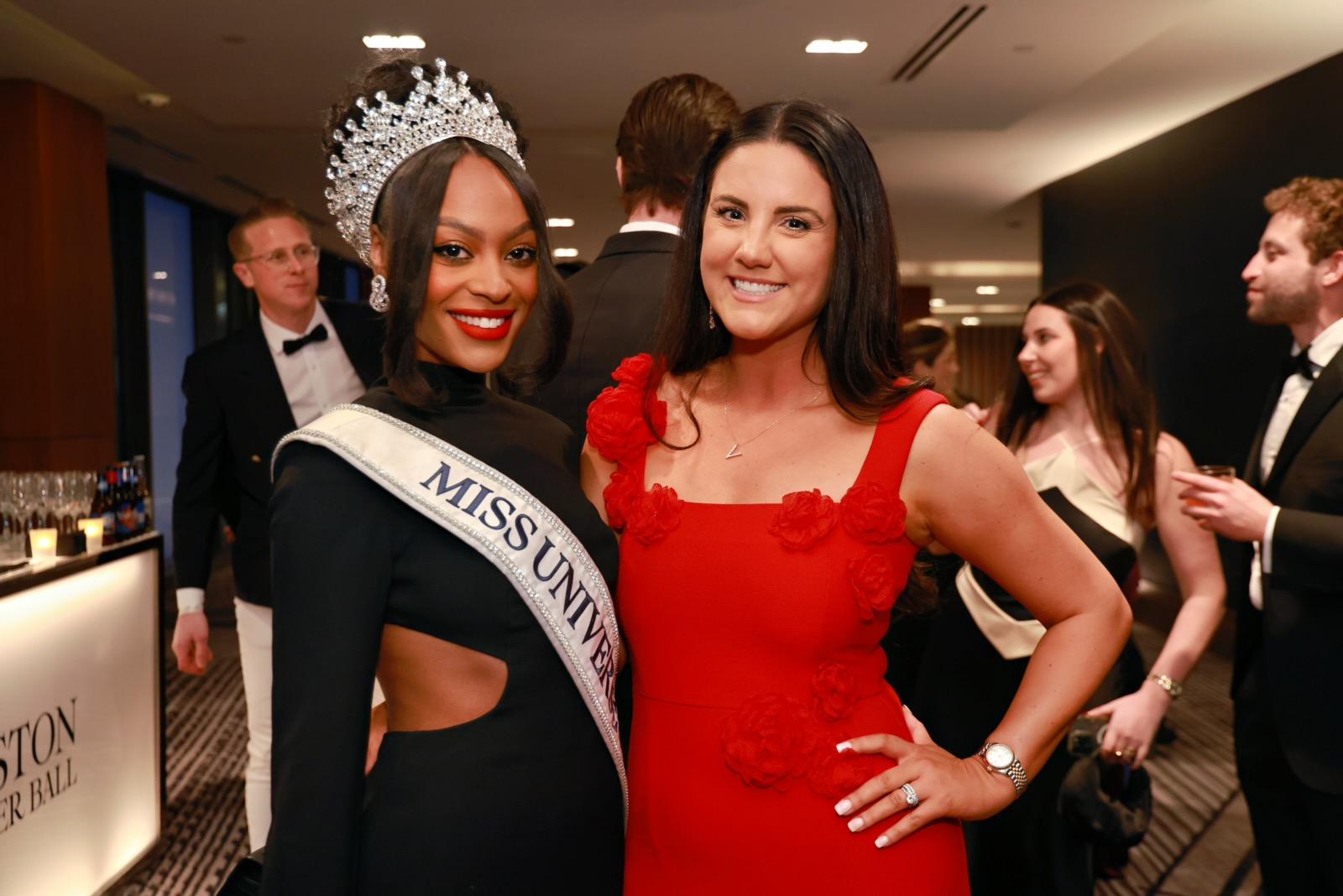 A woman wearing a black dress with cutouts, a large jeweled crown, and a "MISS UNIVERSE" sash stands next to another woman in a red dress adorned with fabric flowers. Both women are smiling and posing for the photo. In the background, other people dressed in formal attire are engaged in conversation. The setting appears to be an elegant indoor event.