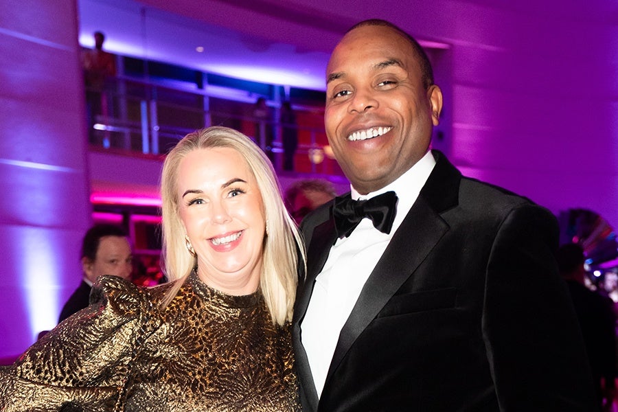 A smiling woman with long blonde hair wearing a shiny gold patterned dress stands next to a smiling man in a black tuxedo with a white shirt and black bow tie. The background features purple and pink lighting with blurred people and architectural elements.