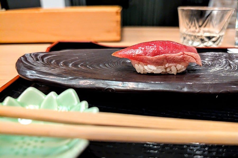 A piece of nigiri sushi with a slice of raw tuna on top of vinegared rice, served on a dark, textured ceramic plate. In the foreground, there are wooden chopsticks resting on a light green ceramic chopstick holder shaped like a leaf. A clear glass with a cut pattern is visible in the background.