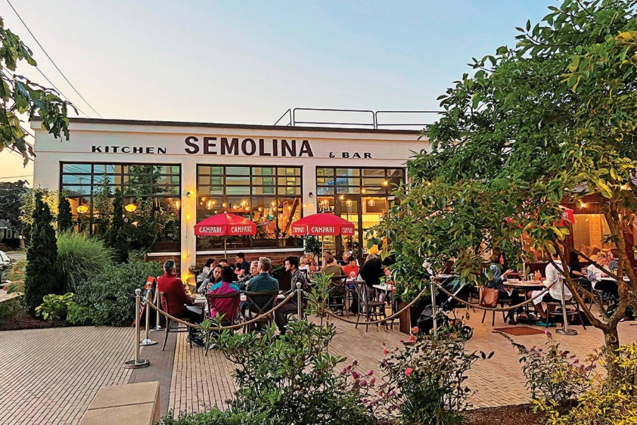 Outdoor dining area of Semolina Kitchen & Bar with several people seated at tables under red Campari umbrellas, surrounded by greenery and trees, during early evening. The building has large windows and warm interior lighting.
