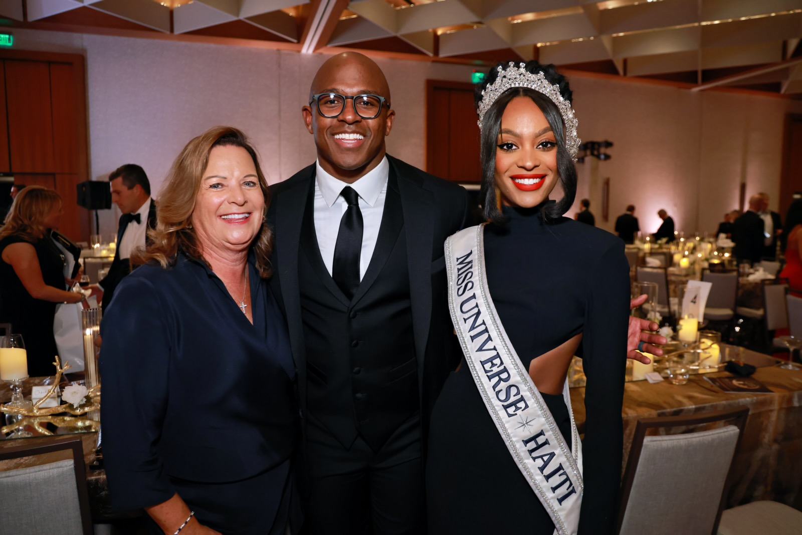 A woman wearing a black dress, a "Miss Universe Haiti" sash, and a jeweled crown stands smiling next to a man in a black suit and tie and a woman in a dark blue outfit. They are indoors at a formal event with tables, candles, and other guests in the background.