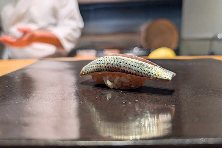 A piece of sushi with a shiny, silver-skinned fish topping on a small mound of rice, placed on a black rectangular plate. The background is blurred, showing a person in a white outfit and some kitchen elements.