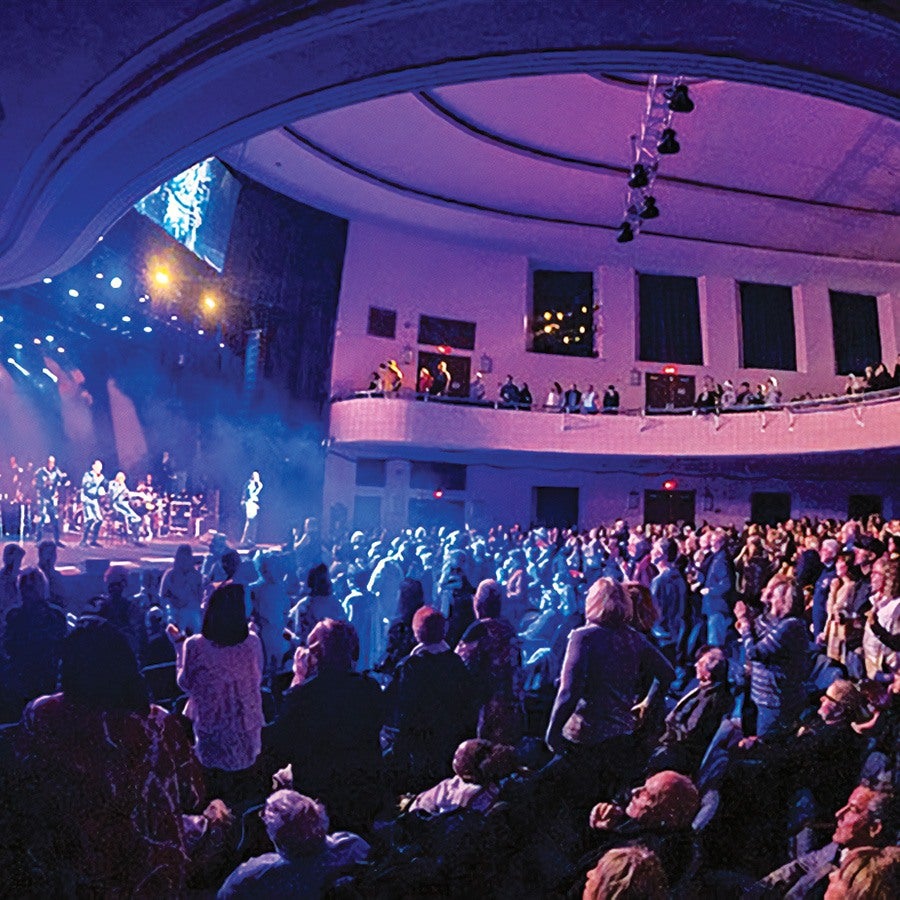 A large indoor concert venue filled with a standing audience watching a live band perform on stage. The stage is illuminated with bright blue and purple lights, and the ceiling features curved architectural details with hanging spotlights. The crowd is densely packed, with some people seated and others standing, facing the stage. A balcony with additional spectators overlooks the main floor.
