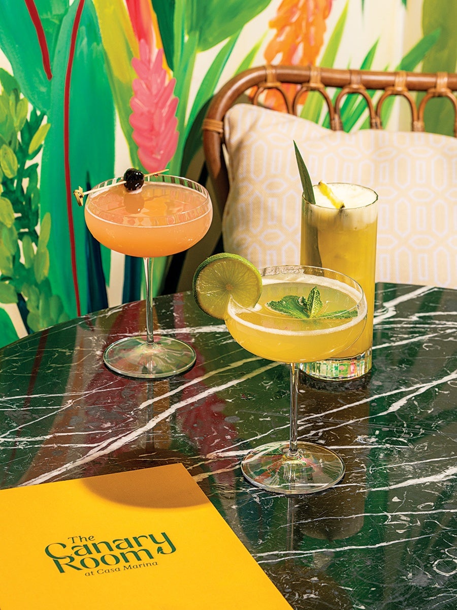 Three colorful cocktails are arranged on a dark green marble table. One cocktail is in a coupe glass garnished with a lime wheel and mint leaves, another is a peach-colored drink in a coupe glass with a cherry garnish, and the third is a tall yellow drink garnished with a pineapple leaf and a pineapple wedge. A yellow menu with "The Canary Room at Casa Marina" printed on it is also on the table. The background features a rattan chair with a patterned cushion and a vibrant tropical mural with green leaves and pink and orange flowers.
