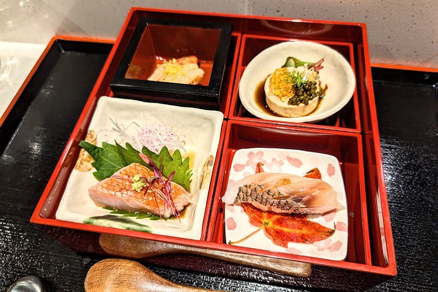 A traditional Japanese bento box with four compartments, each containing different dishes. The top left compartment has a small portion of shrimp with garnish. The top right compartment holds a round tofu or egg dish topped with green caviar and sauce. The bottom left compartment features a piece of grilled salmon on green leaves with a small amount of wasabi and microgreens. The bottom right compartment contains a piece of fish sashimi on a decorative plate with a red maple leaf design. A wooden spoon rests in front of the bento box on a black tray.