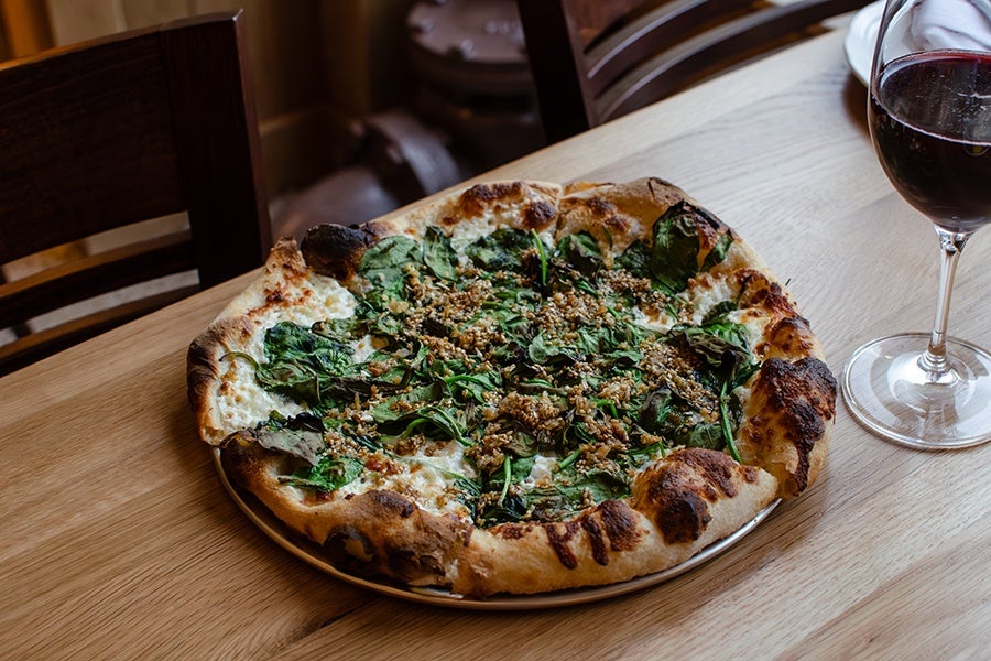 A pizza with a golden-brown crust topped with spinach and a sprinkle of nuts or seeds, placed on a wooden table. Next to the pizza is a glass of red wine.