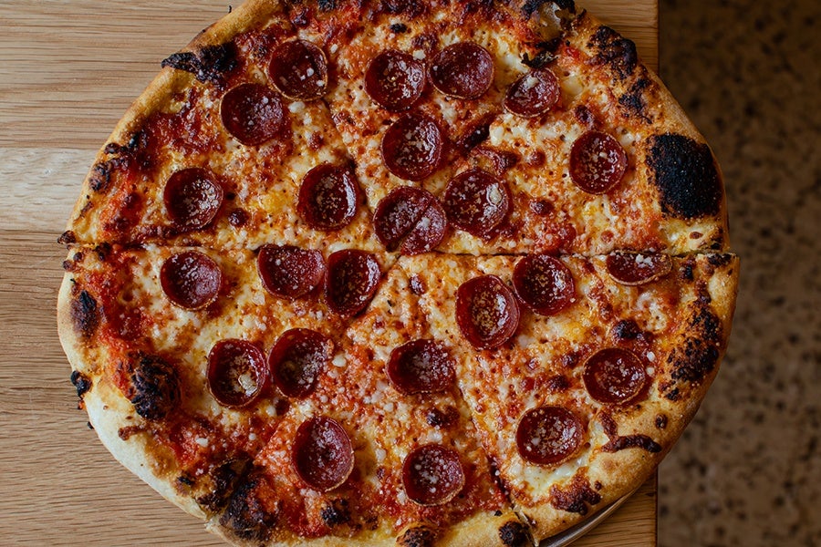 Pepperoni pizza with a golden-brown crust, slightly charred in spots, topped with melted cheese and evenly distributed pepperoni slices, placed on a wooden surface.