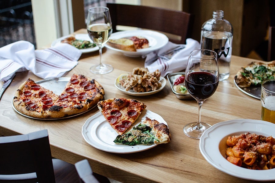A wooden table set with various dishes including a pepperoni pizza with a slice missing, a white plate with two pizza slices (pepperoni and spinach), a bowl of fried calamari with a lemon wedge, a plate of pasta with tomato sauce, and a partially eaten spinach pizza. Drinks include a glass of white wine, a glass of red wine, a glass of beer, and a bottle of water. White napkins with red stripes are folded on the table.