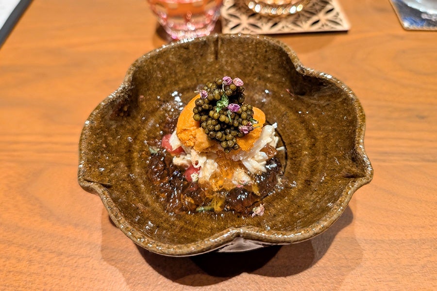 A gourmet dish served in a rustic, brown, flower-shaped ceramic bowl. The dish features a layered presentation with a base of finely chopped ingredients, topped with a bright orange sea urchin (uni), a generous dollop of black caviar, and garnished with small purple edible flowers. The bowl is placed on a wooden table.